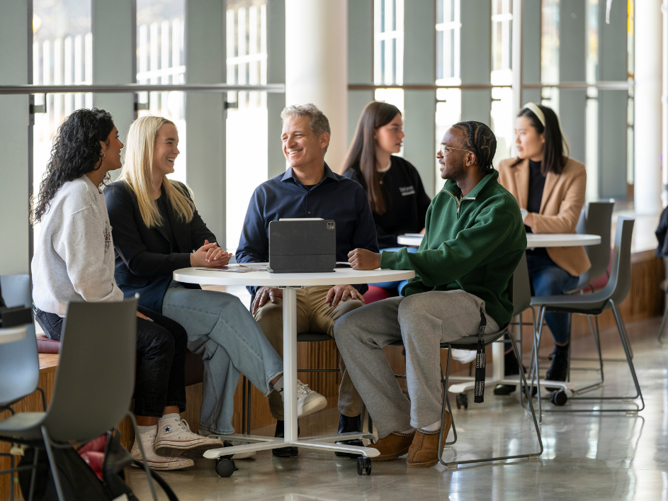 Group of 3 students and faculty member sitting at a table in a common area working together