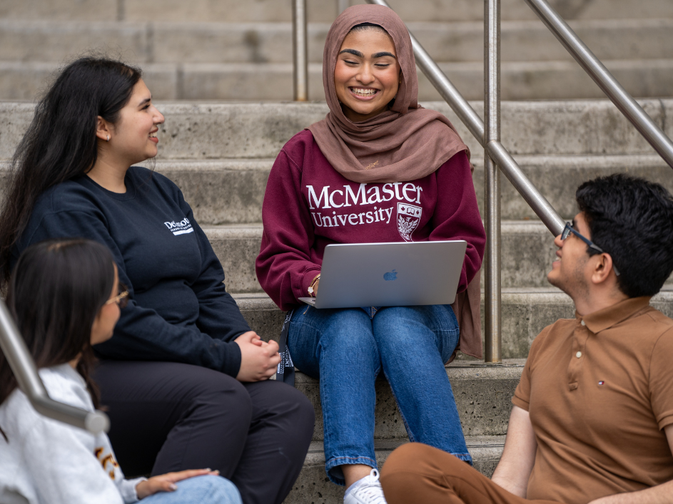 Students sitting outdoors on concrete stairs, facing inwards towards student wearing a hijab with a laptop computer in her lap, smiling warmly